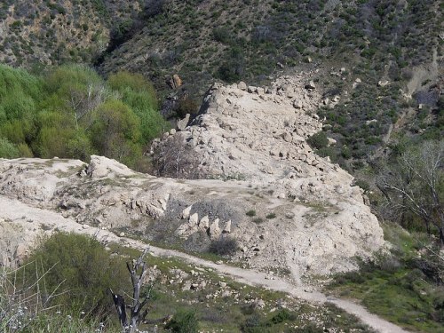 Rocky terrain with a steep slope, surrounded by greenery and shrubs in a hilly landscape.