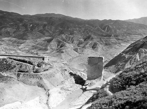 Black and white photo of a large dam in a mountainous landscape, showcasing rugged terrain and distant hills.