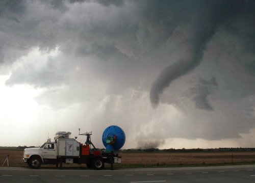 A tornado forms in the distance near a storm-chasing vehicle parked on a rural road. Dark clouds loom overhead.