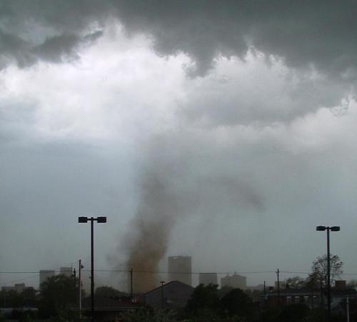 A dark, cloudy sky with a visible tornado forming in the distance, near urban buildings and power lines.