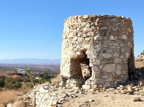 Stone ruins of a circular structure on a hillside, overlooking a valley and distant mountains under a clear blue sky.