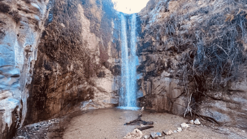 A serene waterfall cascading down rocky cliffs into a sandy area, surrounded by dry vegetation.