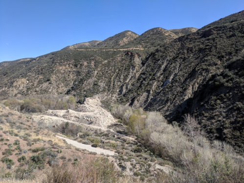 A scenic view of rugged mountains and a dry riverbed, with sparse vegetation under a clear blue sky.