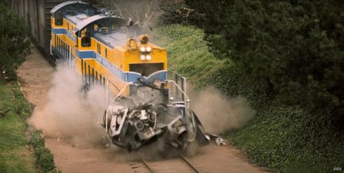 A train collides with a wrecked vehicle, creating a cloud of dust and debris on the tracks.