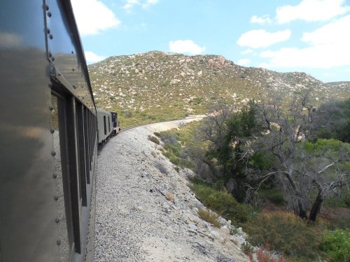 A train winds along a rocky path through green hills under a blue sky with scattered clouds.