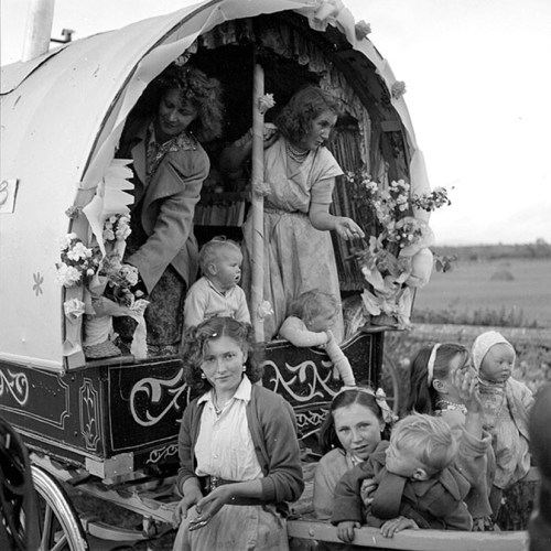 A vintage photo of women and children around a decorated horse-drawn caravan, with flowers and playful expressions.