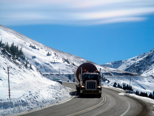 A large truck navigates a winding road through snowy mountains under a clear blue sky.