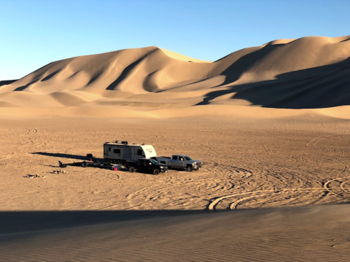 A camper and truck parked in a vast desert landscape with rolling sand dunes under a clear blue sky.