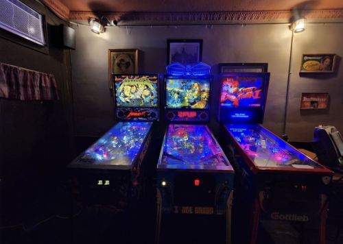 Three illuminated pinball machines in a dimly lit room, featuring vibrant artwork and colorful lights.