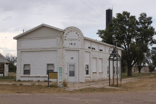 Historic white building with "Bank of Baca County 1912" sign, surrounded by grass and trees, under a cloudy sky.