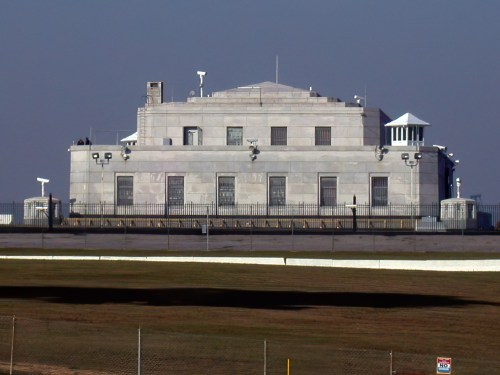 A large, modern building with a flat roof, surrounded by a fence and surveillance equipment, set against a clear sky.