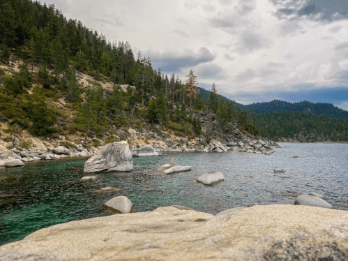 A serene lake surrounded by rocky shores and lush green trees under a cloudy sky.