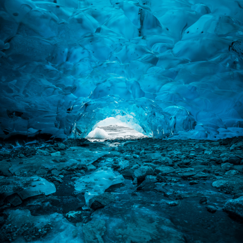 A stunning ice cave with vibrant blue walls and a bright light at the entrance, surrounded by rocky terrain.