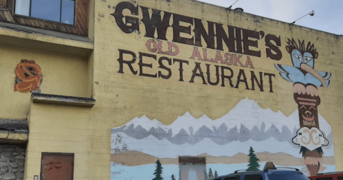 Mural of "Gwennie's Old Alaska Restaurant" with a totem and mountain scenery on a building exterior.