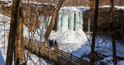A frozen waterfall surrounded by snow-covered trees, with a wooden bridge and people admiring the view.