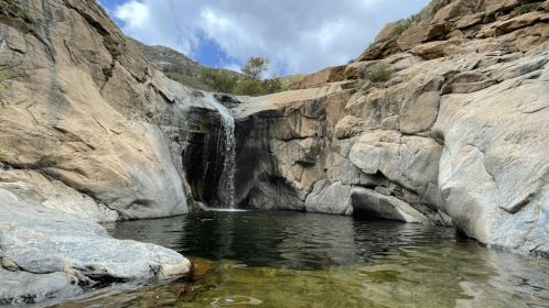 A serene waterfall cascades over rocky cliffs into a calm pool, surrounded by rugged terrain and cloudy skies.