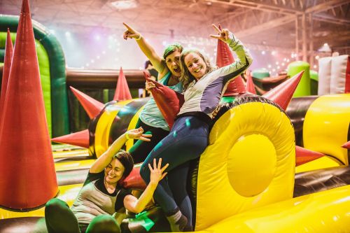 Three friends playfully pose on a colorful inflatable obstacle course, surrounded by bright spikes and festive lights.