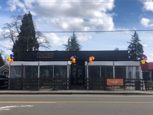 A restaurant with a black exterior, white tents, and colorful balloons outside, set against a blue sky with clouds.