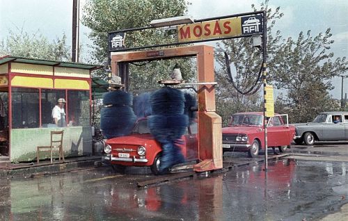 A vintage car wash with colorful brushes, a small car being washed, and a person in a booth nearby.