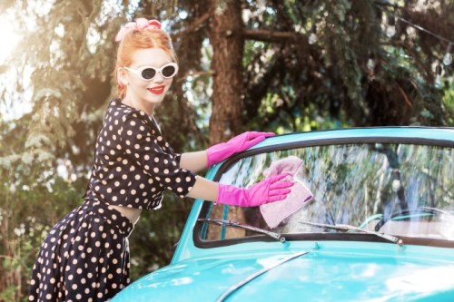 A woman in a polka dot dress and pink gloves cleans the windshield of a vintage turquoise car.