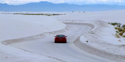 A red car drives along a winding road through white sand dunes with mountains in the background.
