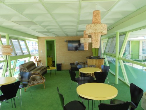 A modern lounge area with green walls, yellow tables, black chairs, and unique light fixtures. Large windows provide natural light.