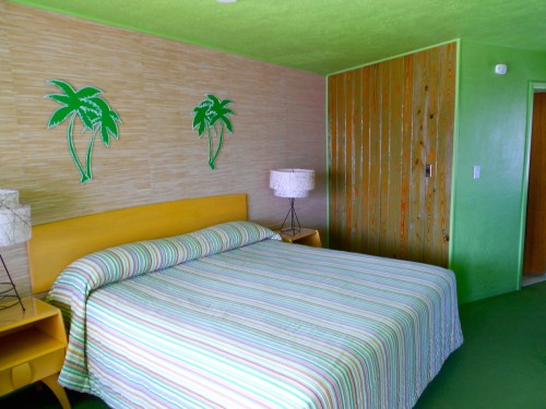 A brightly colored bedroom with a green wall, striped bedspread, and palm tree decorations. Two lamps on nightstands.
