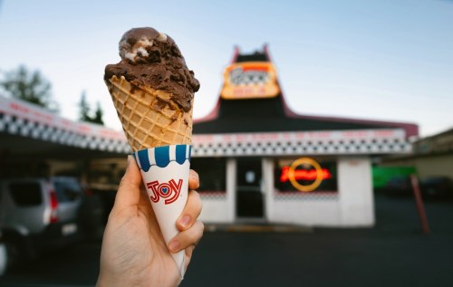 A hand holds a chocolate ice cream cone in front of a retro ice cream shop with a neon sign.