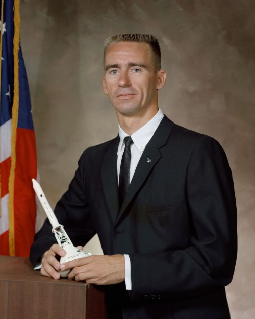 A man in a suit stands beside a model rocket, with an American flag in the background.