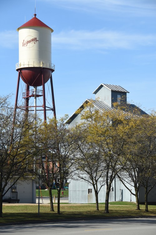 A red water tower labeled "Harmony" next to a gray barn, surrounded by trees with autumn leaves under a clear blue sky.