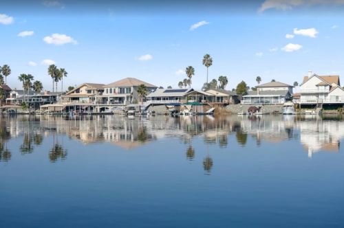 A serene waterfront scene with houses and palm trees reflected in calm water under a clear blue sky.