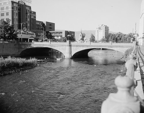 A black and white photo of a bridge over a river, with buildings and trees in the background.