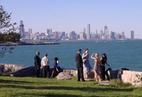 A group of people in formal attire near a lake, with a city skyline in the background on a sunny day.