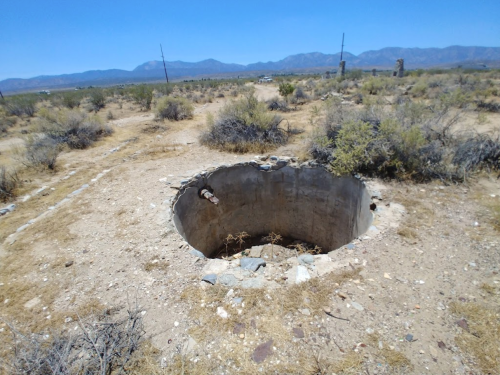 A circular, concrete well surrounded by dry, barren land and sparse vegetation under a clear blue sky.