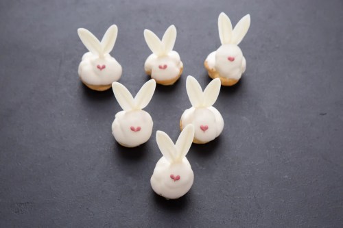 Six cute bunny-shaped desserts with white frosting, pink hearts, and white chocolate ears on a dark surface.