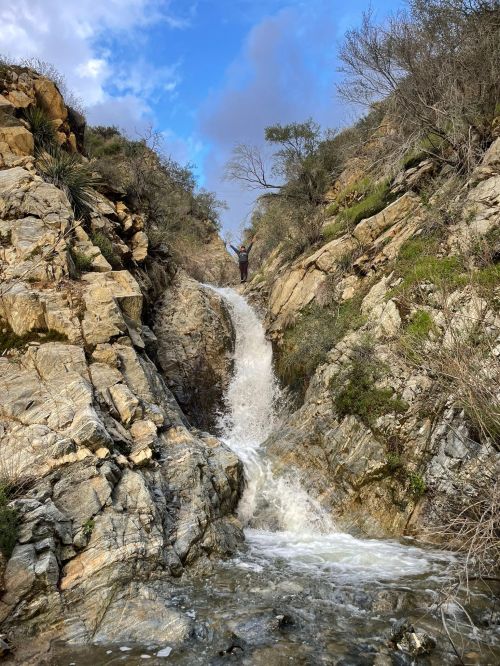 A waterfall cascades down rocky terrain, surrounded by greenery and a blue sky with scattered clouds.