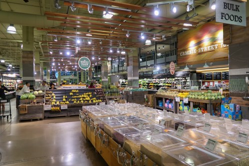 A grocery store interior featuring fresh produce, bulk food bins, and shoppers browsing the aisles.