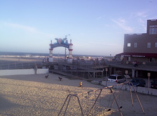 A beach scene with a boardwalk entrance, swings in the foreground, and people walking along the shore at sunset.