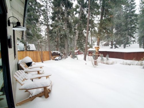 A snowy outdoor scene with wooden chairs, surrounded by tall trees and a fence, creating a serene winter atmosphere.