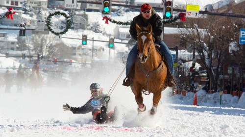 A man on a horse races down a snowy street while a person on a sled is being pulled behind, creating a dynamic winter scene.