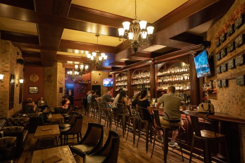 A cozy bar interior with patrons seated at the counter, warm lighting, and shelves filled with bottles.