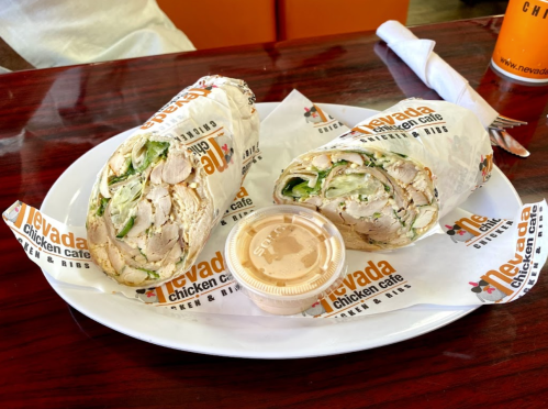 A plate with two halves of a chicken wrap and a small container of sauce, branded with Nevada Chicken Cafe logos.