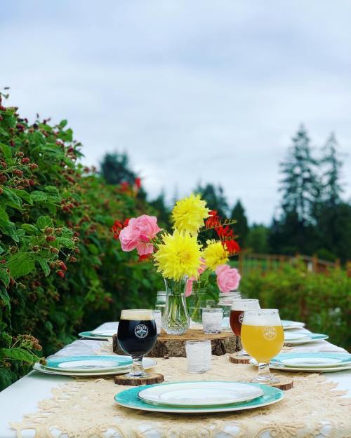 A beautifully set outdoor table with flowers, glasses of beer, and a scenic backdrop of greenery and cloudy skies.