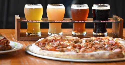 A wooden flight of four craft beers beside a large pizza with various toppings on a table.