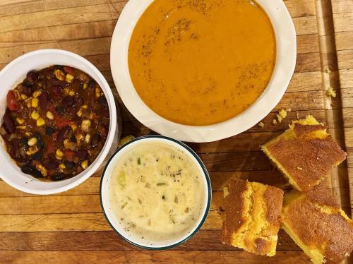 A wooden table with bowls of chili, soup, and a dish of cornbread squares.