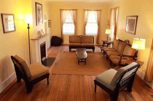 Cozy living room with yellow walls, wooden floors, two armchairs, a sofa, and a coffee table, illuminated by natural light.
