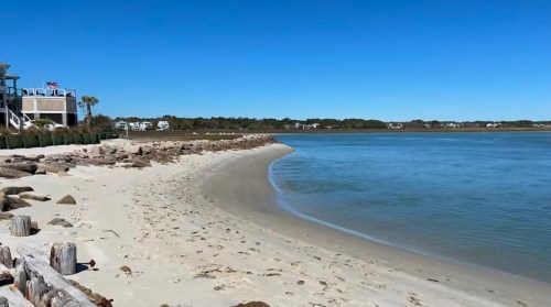 A serene beach scene with a curved shoreline, calm water, and clear blue sky, lined with houses in the distance.