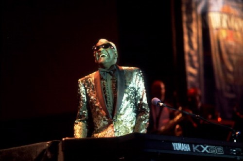 A musician in a shiny gold suit plays a keyboard, smiling and wearing sunglasses on stage.