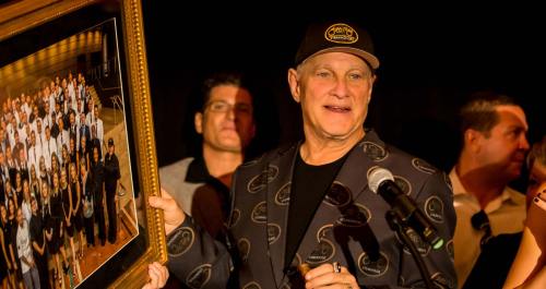 A man in a patterned blazer holds a framed photo of a large group, speaking into a microphone at an event.