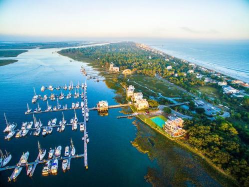 Aerial view of a marina with boats, lush greenery, and a coastline under a clear sky at sunrise.